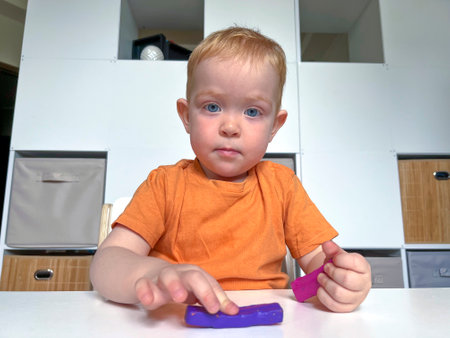 A red-haired boy in an orange T-shirt sculpts blue and lilac plasticine at a table. Child development at home.の写真素材