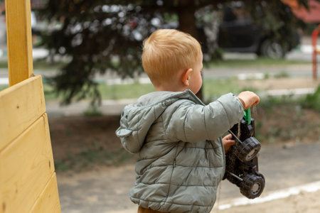 A little red-haired boy in a jacket holds a toy on the playground. Rear view. Outdoor walks.の写真素材