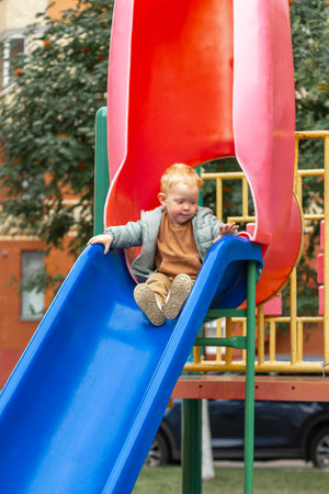A little boy in a sweatshirt and jacket slides down a bright blue slide on a playground in the city. A walk in the fresh air, sincere emotions. Vertical photo.の写真素材