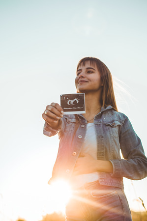 Young Pregnant Woman holding ultrasound photo at Sunset and Embracing her Belly. 4 Month Pregnancy. Maternity Concept. Toned Photo.の写真素材