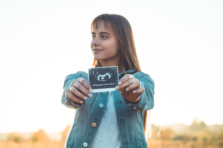 Young Pregnant Woman holding ultrasound photo at Sunset and Embracing her Belly. 4 Month Pregnancy. Maternity Concept. Toned Photo.の写真素材