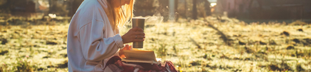 In the morning the girl sits on a wooden bench in the mountains in nature, reads a book and drinks hot tea from a thermo cup. Concepts Reading in nature. Outdoor trainingの写真素材