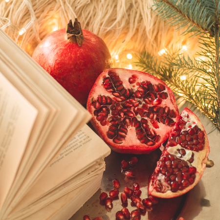 Cozy winter morning at home. Book and Pomegranate in a winter composition, Christmas trees, cones. Warm pillows and garlands on the background. Symbol of New Year and Christmas. Reading timeの写真素材