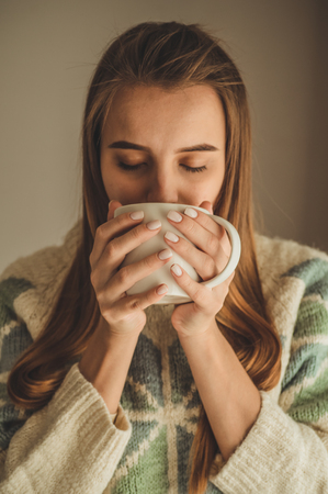 Cozy home. Woman with cup of hot drink  by the window. Looking at window and drink tea. Good morning with tea. Pretty young girl relaxing. Happy concept.の写真素材