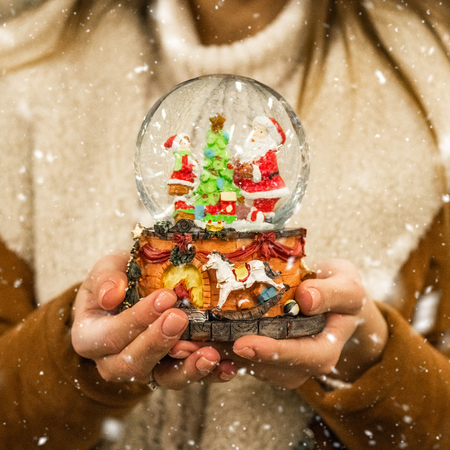 Girl in warm coat holding glass ball with firtrees, house and artificial snow in a mall at the Christmas Fair. Winter mood.の写真素材
