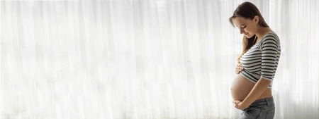 Happy Attractive pregnant woman standing near the window and holding her belly. Concepts of...