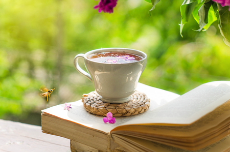 Book, glasses, cup of tea and lilac on a wooden window. The bee flies beautifully over the lilac petals. Fragrant tea in the garden. Romantic concept. Vintage styleの写真素材