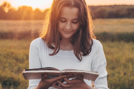 Christian teenage girl holds book in her hands. Reading the book in a field during beautiful sunset. The girl sitting on a grass, reading a book. Rest and readingの写真素材
