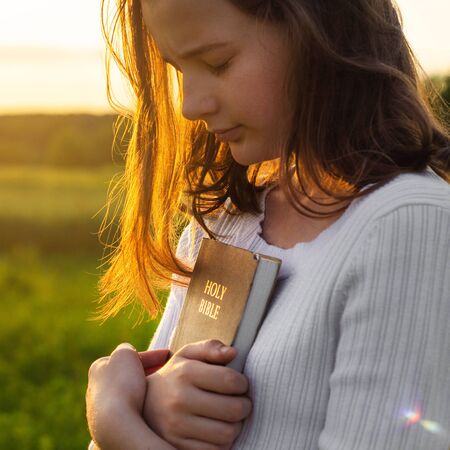 Christian teenage girl holds bible in her hands. Reading the Holy Bible in a field during beautiful...
