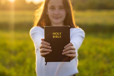 Christian teenage girl holds bible in her hands. Reading the Holy Bible in a field during beautiful sunset. Concept for faith, spirituality and religion. Peace, hopeの写真素材