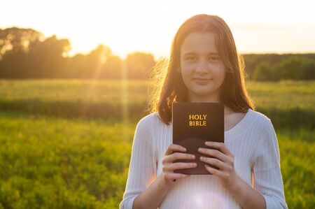 Christian teenage girl holds bible in her hands. Reading the Holy Bible in a field during beautiful sunset. Concept for faith, spirituality and religion. Peace, hopeの写真素材