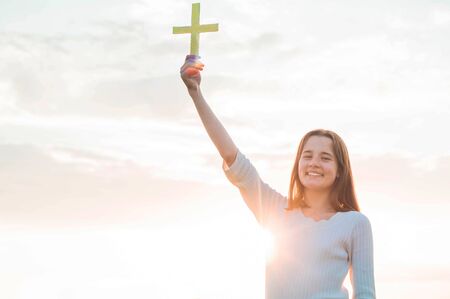 Teenager Girl holding a the cross in hand during beautiful sunset. Hands folded in prayer concept for faith, spirituality and religion. Peace, hope, dreams conceptの写真素材
