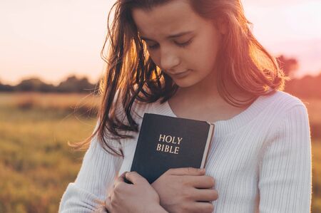 Christian teenage girl holds bible in her hands. Reading the Holy Bible in a field during beautiful sunset. Concept for faith, spirituality and religion. Peace, hopeの素材 [FY310127901789]