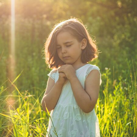 Little Girl closed her eyes, praying in a field during beautiful sunset. Hands folded in prayer concept for faith, spirituality and religion. Peace, hope, dreams conceptの写真素材