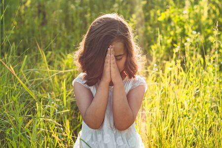 Little Girl closed her eyes, praying in a field during beautiful sunset. Hands folded in prayer concept for faith, spirituality and religion. Peace, hope, dreams conceptの写真素材