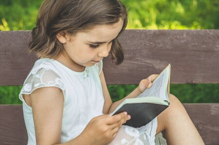 Little Girl holds book in her hands. Reading the book in in outdoors. The girl on sitting on a bench, reading a book. Rest and readingの写真素材