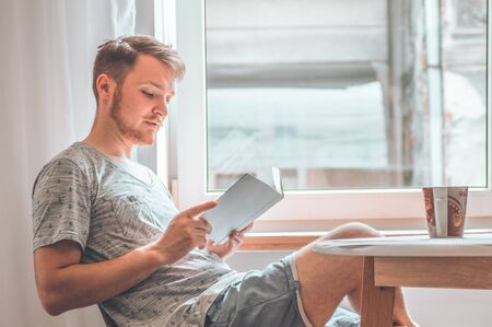 Attractive young man is reading a book at home. Thoughtful guy reading important book. Reading and developmentの写真素材