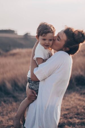 Young woman kisses her little son at sunny in the meadow in sunset. Young mother holding her baby. Mother and  little son having good time in the nature. Color toned image. Mothers Day. Family Conceptの写真素材