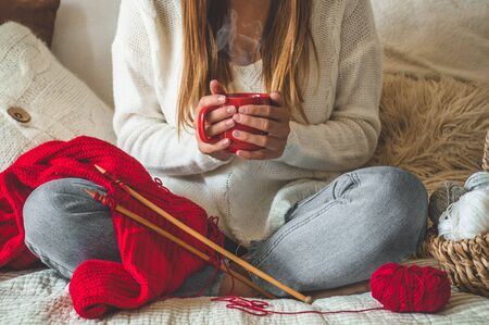 Beautiful girl knits a warm sweater with a hot cup of tea on the bed. Knitting as a hobby. Accessories for knitting.の写真素材