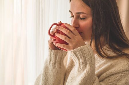 Woman with cup of hot drink by the window. Looking at window and drink tea. Good morning with tea. Autumn Winter Time. Cozy home.の写真素材