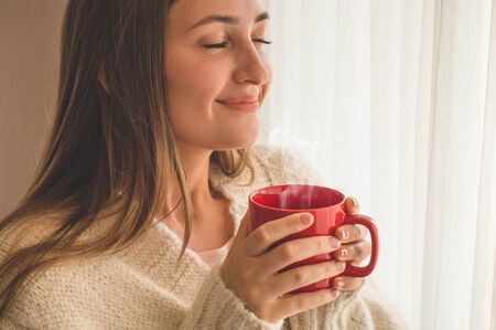 Woman with cup of hot drink by the window. Looking at window and drink tea. Good morning with tea. Autumn Winter Time. Cozy home.の写真素材