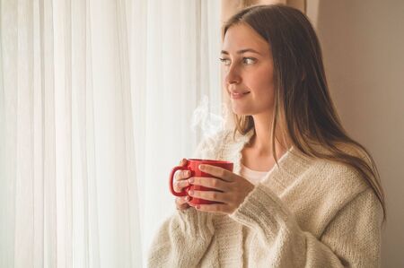 Woman with cup of hot drink by the window. Looking at window and drink tea. Good morning with tea. Autumn Winter Time. Cozy home.の写真素材