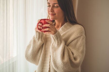 Woman with cup of hot drink by the window. Looking at window and drink tea. Good morning with tea. Autumn Winter Time. Cozy home.の写真素材