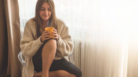 Woman with cup of hot drink by the window. Looking at window and drink tea. Good morning with tea. Autumn Winter Time. Cozy home.の写真素材