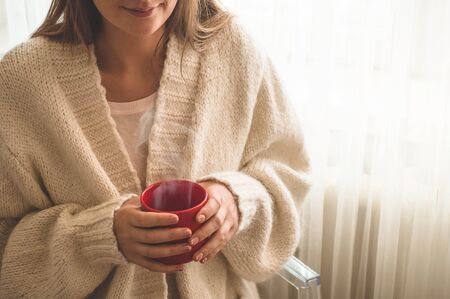 Woman with cup of hot drink by the window. Looking at window and drink tea. Good morning with tea. Autumn Winter Time. Cozy home.の写真素材