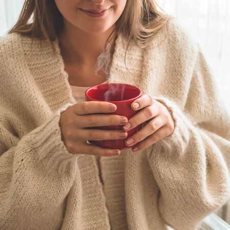 Woman with cup of hot drink by the window. Looking at window and drink tea. Good morning with tea. Autumn Winter Time. Cozy home.の写真素材