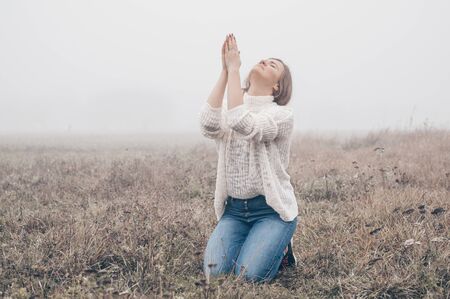 Girl closed her eyes on the knees, praying in a field during beautiful fog. Hands folded in prayer concept for faith, spirituality and religion. Peace, hope, dreams conceptの写真素材