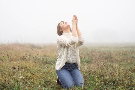 Girl closed her eyes on the knees, praying in a field during beautiful fog. Hands folded in prayer concept for faith, spirituality and religion. Peace, hope, dreams conceptの写真素材