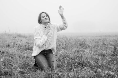 Girl closed her eyes on the knees, praying in a field during beautiful fog. Hands folded in prayer concept for faith, spirituality and religion. Peace, hope, dreams conceptの写真素材