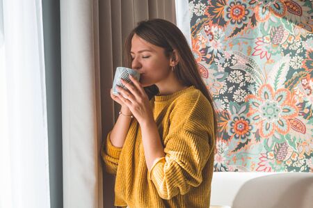 Woman with cup of hot drink by the window. Looking at window and drink tea. Good morning with tea. Autumn Winter Time. Cozy home.の写真素材