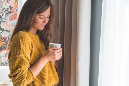 Woman with cup of hot drink by the window. Looking at window and drink tea. Good morning with tea. Autumn Winter Time. Cozy home.の写真素材
