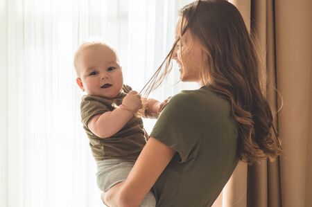 Home portrait of a baby boy with mother near the window. Mom holding and kissing her child. Mothers...