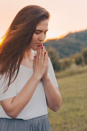 Portrait of a single woman praying and looking down at sunset. Hands folded in prayer concept for faith, spirituality and religion. Peace, hope, dreams conceptの写真素材
