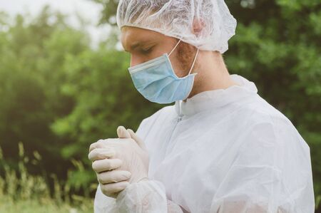 Young man in protective medical mask and latex gloves praying outdoors. Concept of coronavirus, pandemic, prayer, appeal to God, religionの写真素材
