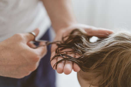 Father cuts her son hair in the room. Family during quarantine, Scissors and a comb in male hands. Boy with a new haircut.の写真素材