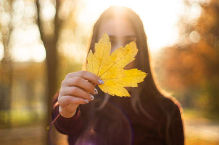 Girl holding Yellow leaf in hand. Beautiful autumn model with waving glow hair. Portrait of romantic...