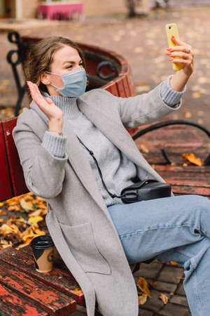 Girl in protective mask using phone for video calls to friends in the park. Technology concept.