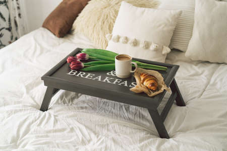Tray with croissant, cup of coffee and tulip in white clean bed.