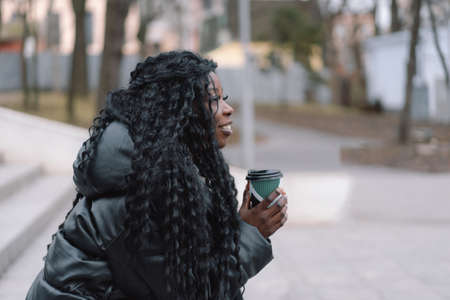 Cheerful black skinned girl keeping paper cup of hot coffee walking in city park
