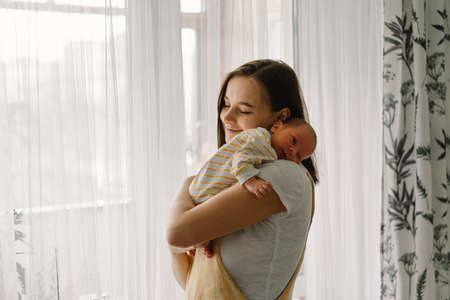 Mother playing with newborn baby son at home near the window. Happy infant and mom. Isolation during...
