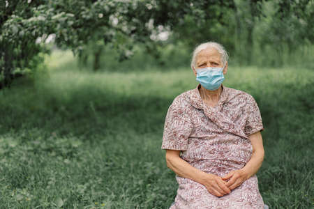 Portrait of a gray-haired sad old woman alone wearing protective face mask while during quarantine.