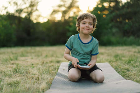Little boy sitting on a sports mat in a outdoors and using mobile phone. Concept of lifestyle