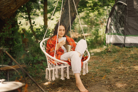 Girl wrapped in plaid drinking tea and use phone in a hanging chair outdoors. People use technologyの写真素材