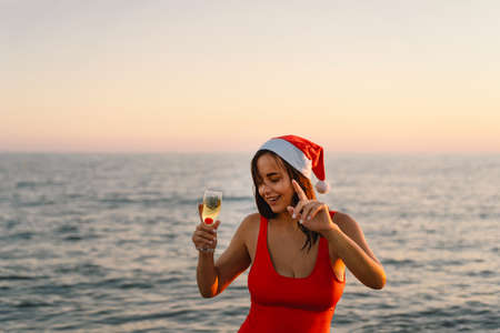 Young beautiful woman in a Santa hat with a glass of champagne in her hands walk on the beach.の写真素材