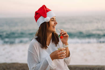 Young beautiful woman in a Santa hat with a glass of cocktail in her hands walk on the beach.の写真素材