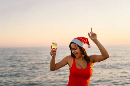 Young beautiful woman in a Santa hat with a glass of champagne in her hands walk on the beach.の写真素材